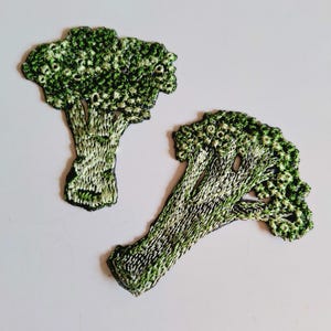 May include: Two embroidered broccoli patches. The patches feature detailed stitching in shades of green and white, mimicking the texture of broccoli florets and stems. The patches are designed to be sewn onto fabric.