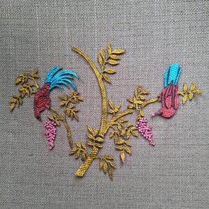 Pode incluir: Um emblema bordado apresentando um galho de árvore dourado com folhas e dois cachos pendentes de flores cor-de-rosa. Dois pássaros com corpos vermelhos e penas de cauda azul brilhante estão pousados nos galhos, voltados para o centro.