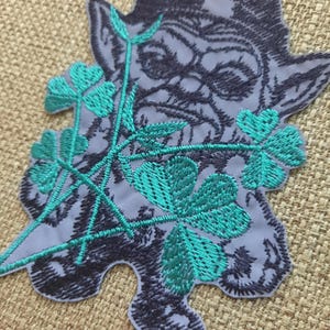 May include: A black and gray embroidered patch of a goblin's face with green shamrocks.