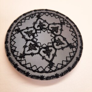 May include: A round, gray fabric patch with a black embroidered design. The design features a five-pointed star with intricate floral details.