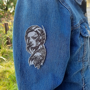 Pode incluir: Um patch bordado em preto e branco com um retrato estilizado de uma pessoa com cabelo comprido, preso a uma jaqueta jeans azul. O patch é um design artístico, adequado para roupas ou acessórios.