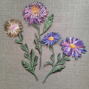 Puede incluir: Tres parches bordados de flores con costuras detalladas. Una flor tiene pétalos naranjas y marrones, mientras que las otras dos muestran pétalos vibrantes de color púrpura, rosa y azul con centros amarillos. Cada flor tiene un tallo y hojas verdes.