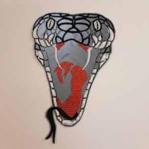 Puede incluir: Un parche bordado de la cabeza de una serpiente, con un diseño gris y negro, una boca roja abierta y una lengua bifurcada negra.