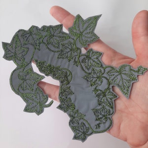 Peut inclure: Un patch en tissu gris avec des vignes et des feuilles brodées en vert. Le patch a la forme d'une vigne et une surface rugueuse et texturée.