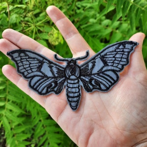 Könnte beinhalten: Ein grauer gestickter Patch eines Nachtfalters mit schwarzen Umrissen. Der Nachtfalter hat eine Flügelspannweite von etwa 15 cm.