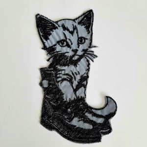 May include: A gray embroidered iron-on patch of a kitten wearing a boot. The kitten is outlined in black thread and has a black boot with a pointed toe.