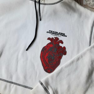 May include: White hoodie with a black drawstring and a red embroidered heart with a black outline. The text "FEARLESS CONNECTION WORLDWIDE/INTL." is printed in black on the hoodie.
