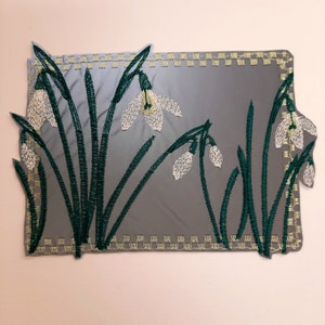 May include: Embroidered white snowdrop flowers with green stems and leaves on a gray background. The design is framed with a yellow stitched border.