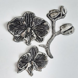 May include: Three black and silver embroidered orchid flower patches. The flowers have a detailed design with a stem and leaves.