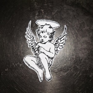 Puede incluir: Un parche bordado blanco con un querubín con alas y un halo. El querubín está sentado con las manos juntas. El parche está sobre una superficie oscura y texturizada.