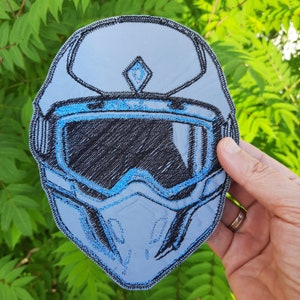 May include: A blue and black embroidered patch of a motorcycle helmet with a visor. The helmet has a diamond shape on the top.