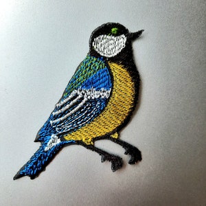 Puede incluir: Parche bordado para planchar de un pájaro azul con detalles amarillos, verdes y blancos. El pájaro está posado en una rama con las alas extendidas.