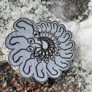 Könnte beinhalten: Ein grauer Stoff-Patch mit einem schwarzen gestickten Design eines Spiralmusters. Das Design besteht aus einer Reihe von sich überlappenden Kreisen und Kurven.