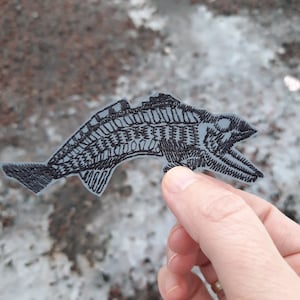 May include: A black embroidered fish patch with a detailed design. The patch is on a light blue background.