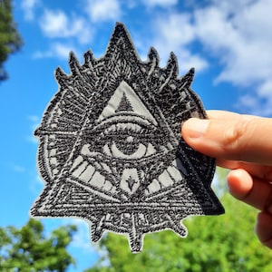 Può includere: Toppa ricamata in bianco e nero di un occhio all'interno di un triangolo con un cerchio attorno. L'occhio guarda direttamente lo spettatore.