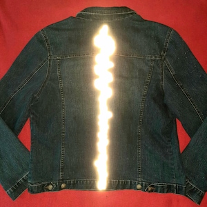 Peut inclure: Une veste en jean bleu foncé avec une longue bande lumineuse blanche verticale au centre du dos. La veste présente un col classique, des manches longues, des coutures apparentes et des boutons métalliques le long de l'ourlet inférieur.