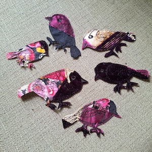May include: Six fabric bird appliqués in shades of pink, purple, black, and gold. The birds feature detailed stitching and varying wing designs. Each bird has a distinct shape and pose, creating a whimsical aesthetic. The appliqués are designed for crafting.
