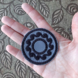 Può includere: Una toppa grigia, circolare, con un disegno ricamato nero. Il disegno presenta un cerchio centrale circondato da una serie di motivi a spirale, simili a onde. La toppa è tenuta in una mano.