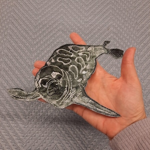 Könnte beinhalten: Ein grau-silberner, gestickter Aufnäher in Form eines Meeresbewohners, möglicherweise ein Wal oder eine Robbe. Der Aufnäher hat aufwendige Details und wird in einer Hand gehalten.