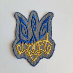 May include: A blue and yellow embroidered patch depicting the Ukrainian coat of arms, the Tryzub, on a gray background.