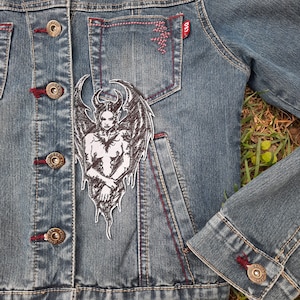 Puede incluir: Una chaqueta vaquera con un parche bordado en blanco y negro de un demonio con alas. El parche está en el lado izquierdo de la chaqueta, cerca del bolsillo.