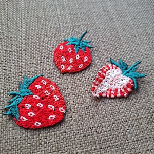 May include: Three embroidered strawberry patches. Two whole strawberries and one sliced in half, all with red bodies, white seeds, and green stems. The patches are on a textured, neutral-colored fabric.