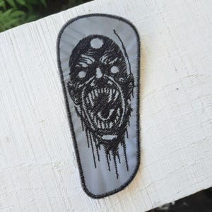 May include: A black and white embroidered patch of a screaming monster's face on a gray background.