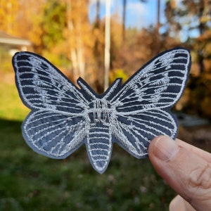 May include: A white embroidered moth design on a black fabric patch. The moth has intricate details and a delicate appearance.