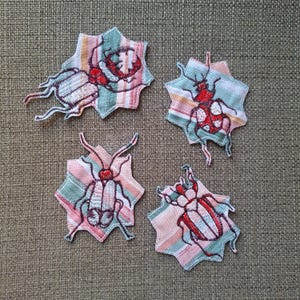 Op de afbeelding: Vier geborduurde insectenpatches. Elke patch heeft een gedetailleerd insectenontwerp in rood, wit en blauw garen, tegen een stervormige achtergrond met streepjespatronen. De patches liggen op een getextureerd, neutraal gekleurd oppervlak.