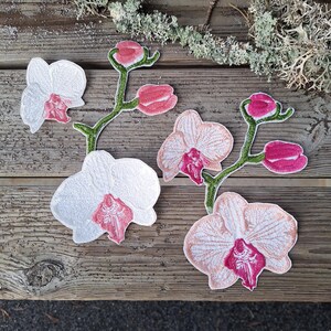 May include: Embroidered orchid patches in white and pink, with green stems and buds. The patches are arranged on a weathered wooden surface, showcasing the delicate details of the floral design. The patches are ideal for fabric crafts.