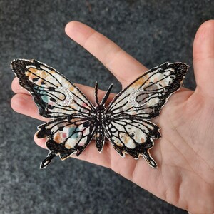 May include: A white and black butterfly fabric patch with a colourful floral pattern. The butterfly has a black outline and is embroidered with white and black details.