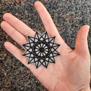 Può includere: Una toppa a forma di stella bianca e nera con un design geometrico. La toppa è fatta di tessuto e ha uno sfondo bianco con un design nero.
