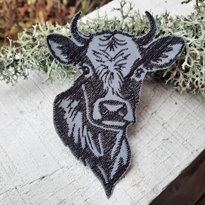Puede incluir: Parche bordado en blanco y negro de la cabeza de una vaca con cuernos. El parche está sobre un fondo gris claro.