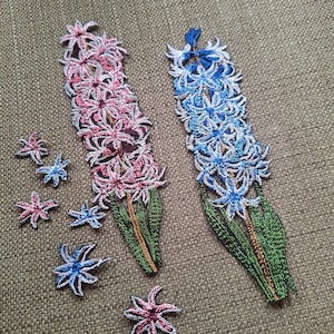 Puede incluir: Dos parches bordados de flores de jacinto, uno en tonos rosas y otro en tonos azules, cada uno con hojas y tallos verdes. Varios parches más pequeños e individuales en forma de estrella con pétalos en colores rosa y azul a juego están dispersos.