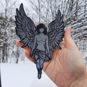 Könnte beinhalten: Ein gestickter Bügeleisen-Aufnäher in Schwarzweiß, der einen Engel mit ausgebreiteten Flügeln zeigt. Der Engel ist in einer Gehpose mit einem fließenden Gewand dargestellt.