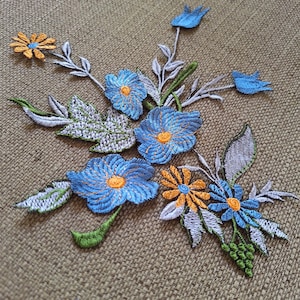 Op de afbeelding: Geborduurde bloemenpatches met blauwe en oranje bloemen, gele centra, groene stelen en witte bladeren. De patches zijn gerangschikt in een boeketachtig ontwerp, perfect voor stoffen versiering of knutselprojecten.