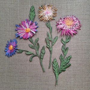 Könnte beinhalten: Drei gestickte Blumenaufnäher mit leuchtenden Blütenblättern in Rosa, Violett, Blau und Orange sowie grünen Stielen und Blättern. Die Designs zeichnen sich durch detaillierte Stickereien und eine strukturierte Fadenführung aus.