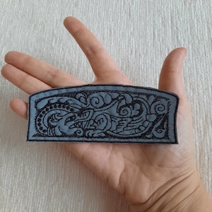 Könnte beinhalten: Ein grauer gestickter Aufnäher mit einem schwarzen Design einer stilisierten Kreatur mit wirbelnden Mustern.