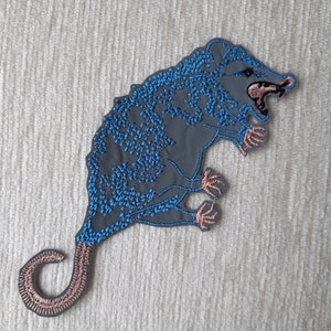 May include: Embroidered iron-on patch of a grey opossum with blue and pink details. The opossum is depicted with its mouth open and its tail curled.