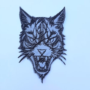 May include: Black and white embroidered patch of a snarling cat's face. The cat has a fierce expression with its mouth open and teeth showing. The patch is perfect for adding a touch of edgy style to clothing, bags, or other accessories.