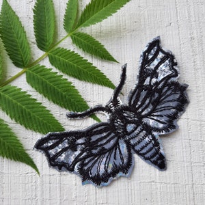 May include: Black and silver embroidered moth patch with iridescent details. The patch is on a white background.