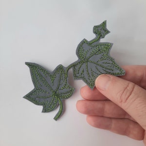 Pode incluir: Uma aplicação de feltro cinza de duas folhas de hera verdes com detalhes bordados.
