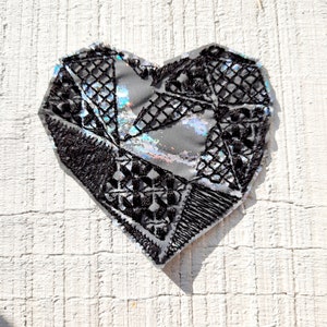 Könnte beinhalten: Ein herzförmiger Patch in Schwarz und Silber mit geometrischem Muster. Der Patch ist aus Stoff und hat eine glänzende, irisierende Oberfläche.