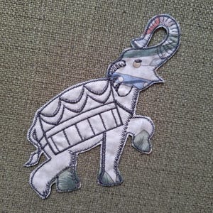 Op de afbeelding: Een geborduurde olifantenpatch met een uniek ontwerp. De olifant is voornamelijk wit met donkerblauwe stiksels die de kenmerken omlijnen. De slurf en de poten van de olifant hebben stoffen accenten met patronen in tinten groen, roze en blauw. De patch staat op een groene achtergrond met textuur.