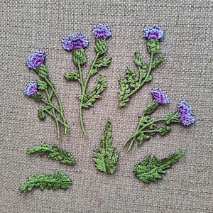 Op de afbeelding: Geborduurde patches met paarse distelbloemen met groene stelen en bladeren. De patches zijn gerangschikt op een getextureerde, neutraal gekleurde achtergrond. De patches zijn gedetailleerd met verschillende tinten paars en groen.