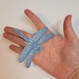 May include: A light blue dragonfly applique with a detailed embroidered design. The applique is made of fabric and has a glittery finish.