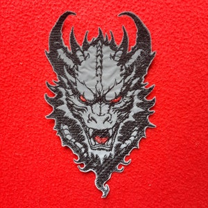 May include: Black and grey embroidered patch of a dragon's head with red eyes. The dragon has sharp teeth and a fierce expression.