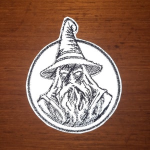 Puede incluir: Parche bordado en blanco y negro de un mago con una larga barba y un sombrero puntiagudo. El parche es circular y tiene un fondo blanco.