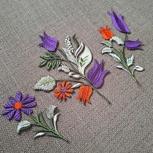 May include: Embroidered floral patches in purple, orange, green, and white. The patches feature detailed stitching of various flowers and leaves, arranged in small bouquets. The background is a textured, neutral-toned fabric.
