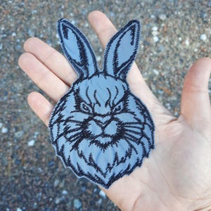 May include: A black and grey embroidered patch of a rabbit's face with a serious expression.